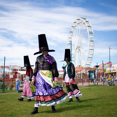 Figures in Welsh costume with fairground behind.