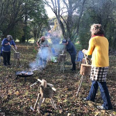 Image of a spoon carving workshop,ran by Graham Beadle with four participants in woodland location chopping and shaping wood,with a smoky firepit in the middle