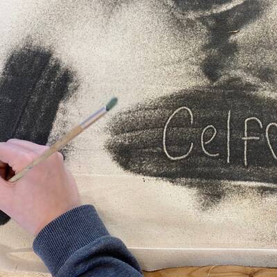 A hand holding a brush and applying charcoal powder. The word "Celf" has been scratched into the charcoal powder.